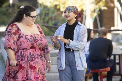  An outdoor scene featuring two individuals engaged in a pleasant conversation. One person wears a vibrant pink floral dress and carries a walking aid, while the other sports a colorful headscarf, a paint-splattered denim shirt, and casual pants. The background shows a sunlit garden with blurred figures at a table, creating a warm, community-oriented atmosphere.