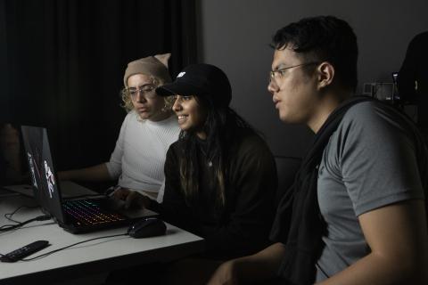 Media Arts Community Three AUArts students collaborating at a laptop with a rainbow-lit keyboard in a dark classroom or studio.