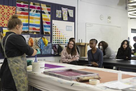 Print Media Community An instructor demonstrates textile printing at a large table while students watch attentively in a classroom decorated with colorful fabric samples and swatches.