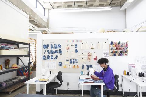 Fibre Community Student sewing at a machine in the AUArts sewing centre, with fabric samples and thread spools displayed on the wall.