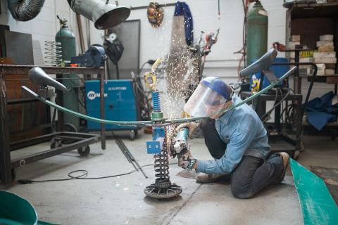 Sculpture Community Student in safety gear uses a grinder on a metal sculpture, creating sparks, inside the AUArts metal shop.