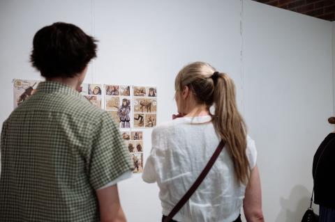 Two people view a wall display of hand-drawn comic panels featuring illustrated characters and scenes.