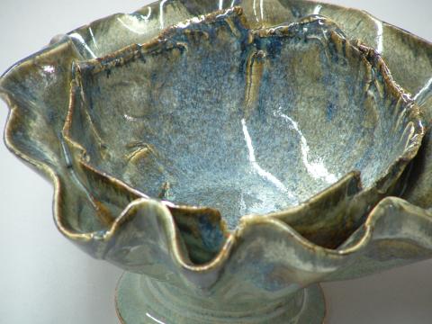 A close up photograph of a ceramic bowl with rippled edges and a narrow base. The glaze is a shimmery pale blue-green