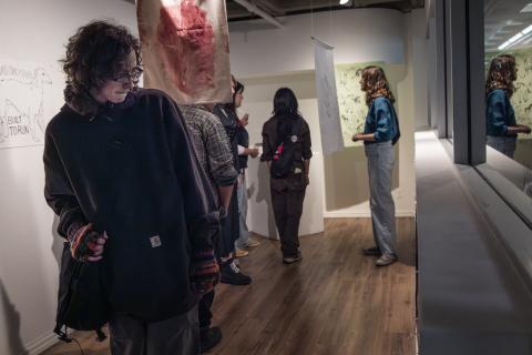 Visitors viewing mixed-media artworks in a contemporary gallery, including hanging textiles and illustrated pieces, during an exhibition event.
