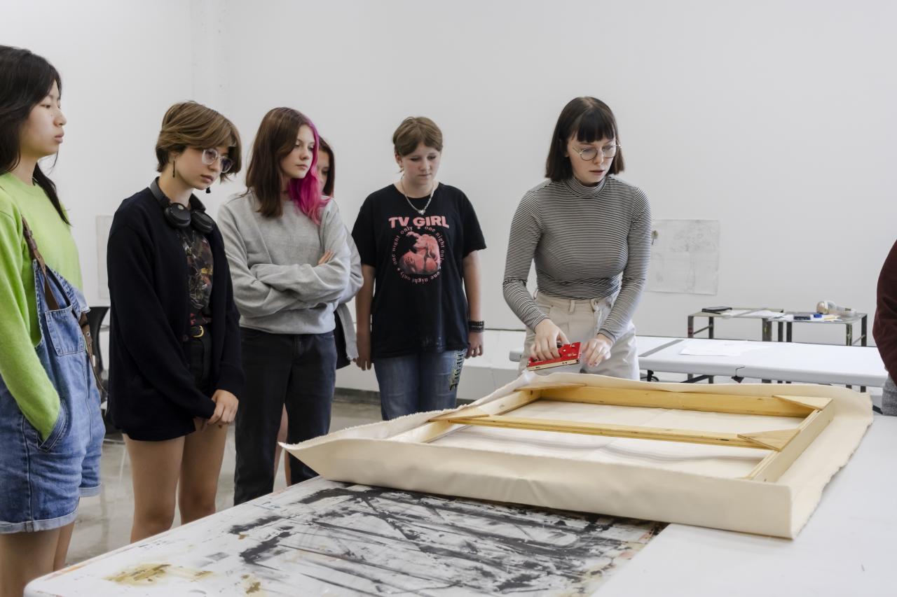 A group of teen painting students observe their instructor guiding them through stretching a canvas