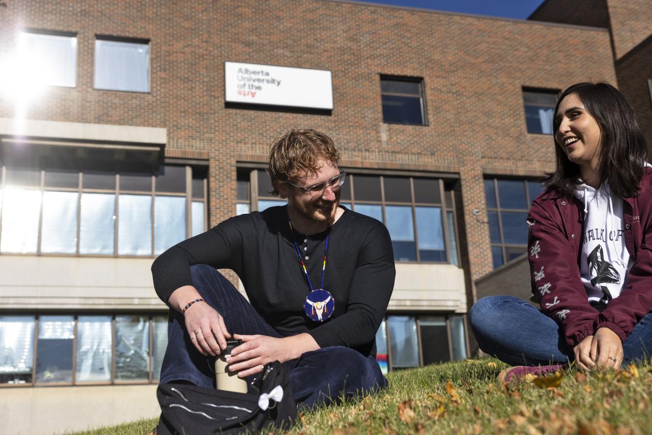 Two students sit on a grassy lawn outside the Alberta University of the Arts building, enjoying a sunny day. The person on the left, wearing glasses, a black shirt, and a colorful beaded necklace, smiles and leans forward with their hands resting on a water bottle. The person on the right, wearing a maroon jacket and hoodie, laughs while sitting cross-legged. Behind them, the university's brick building with large windows and a white sign reading "Alberta University of the Arts".