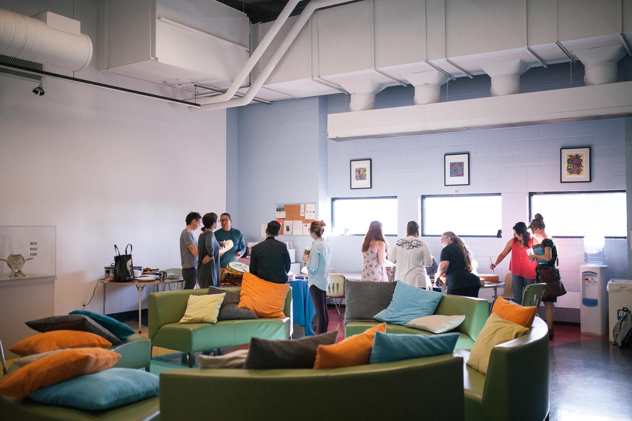 A group of people gathers in a modern, open indoor space with high ceilings and exposed ventilation. The room features light blue and white walls, large windows letting in natural light, and framed colorful artwork. In the foreground, green and gray seating with bright, colorful pillows in shades of orange, blue, and gray creates a cozy, casual atmosphere. The group of individuals stands near a table, engaged in conversation, with some holding cups. The setting appears to be a community or student lounge sp
