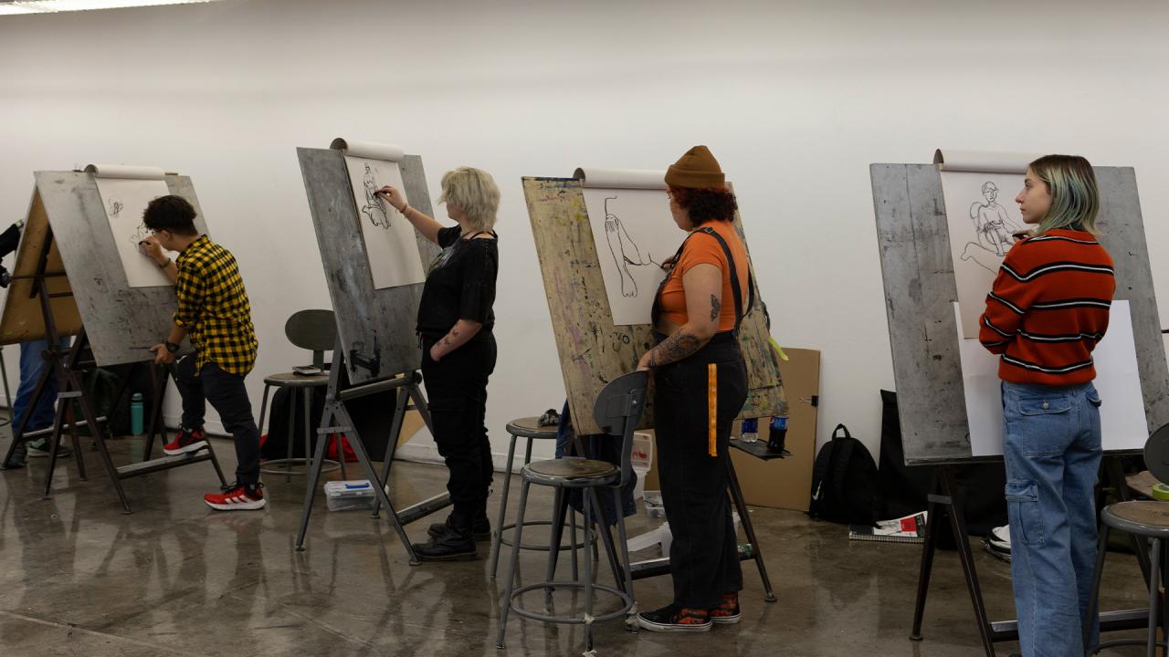 Art students participating in a life drawing class, each standing at an easel and sketching a model. The studio setting features large drawing boards, stools, and an atmosphere of focused creativity as students refine their observational skills.