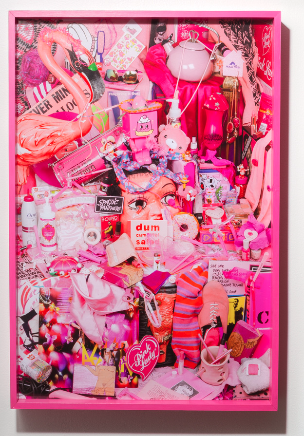 A vibrant, framed collage packed with pink objects, including dolls, flamingos, cosmetics, clothing, and pop culture memorabilia, assembled in a layered, chaotic composition.