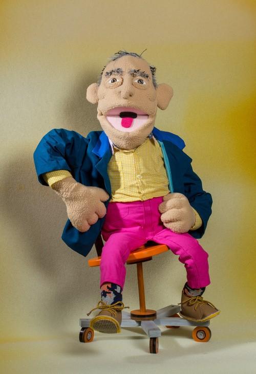 A hand-made puppet of a man with exaggerated facial features, wearing a yellow shirt, pink pants, and a blue jacket, seated on a small rolling chair against a warm yellow backdrop.