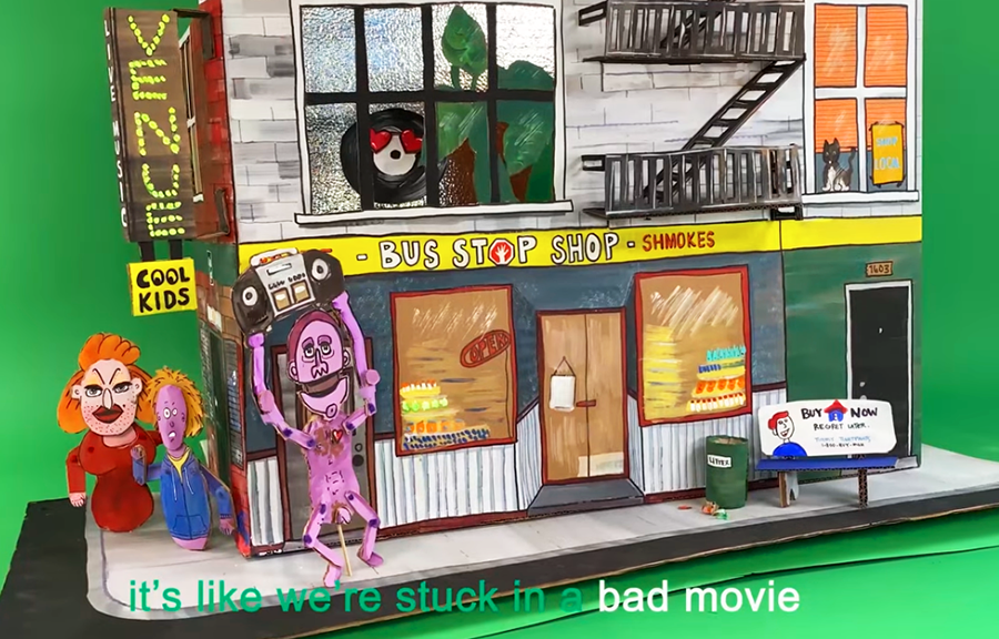 A handcrafted miniature diorama of a colorful urban street scene with cartoonish puppet characters, a storefront named "Bus Stop Shop - Shmokes," and humorous signage, set against a green screen background. A skeleton-like figure holds a boombox while two expressive puppets interact nearby.