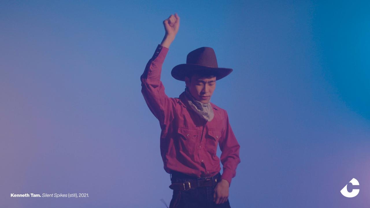 A young man in a cowboy hat, pink shirt, and neckerchief stands with his eyes closed, arm raised in a gesture of expression, against a blue-lit backdrop.