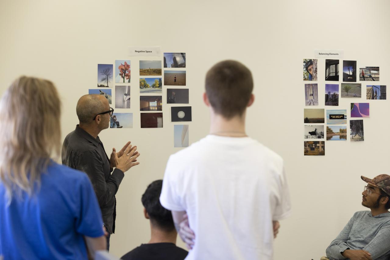 Photography Community Instructor leads a critique as students view photo collages on a classroom wall labeled “Negative Space” and “Balancing Elements.”