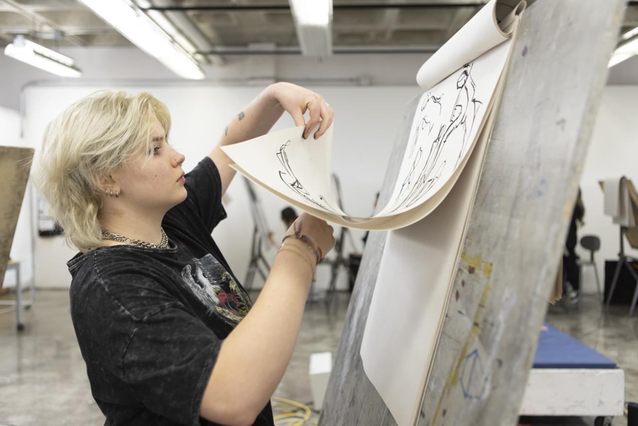 Student flipping through figure drawings on an easel in an AUArts classroom.