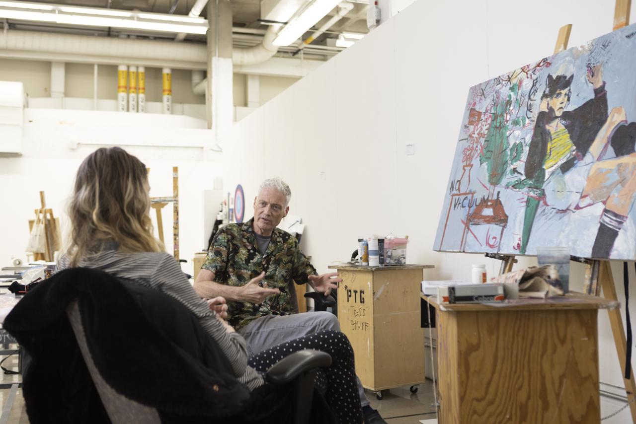 An instructor and student sit in a studio space having a discussion, with a colorful expressive painting on an easel beside them.