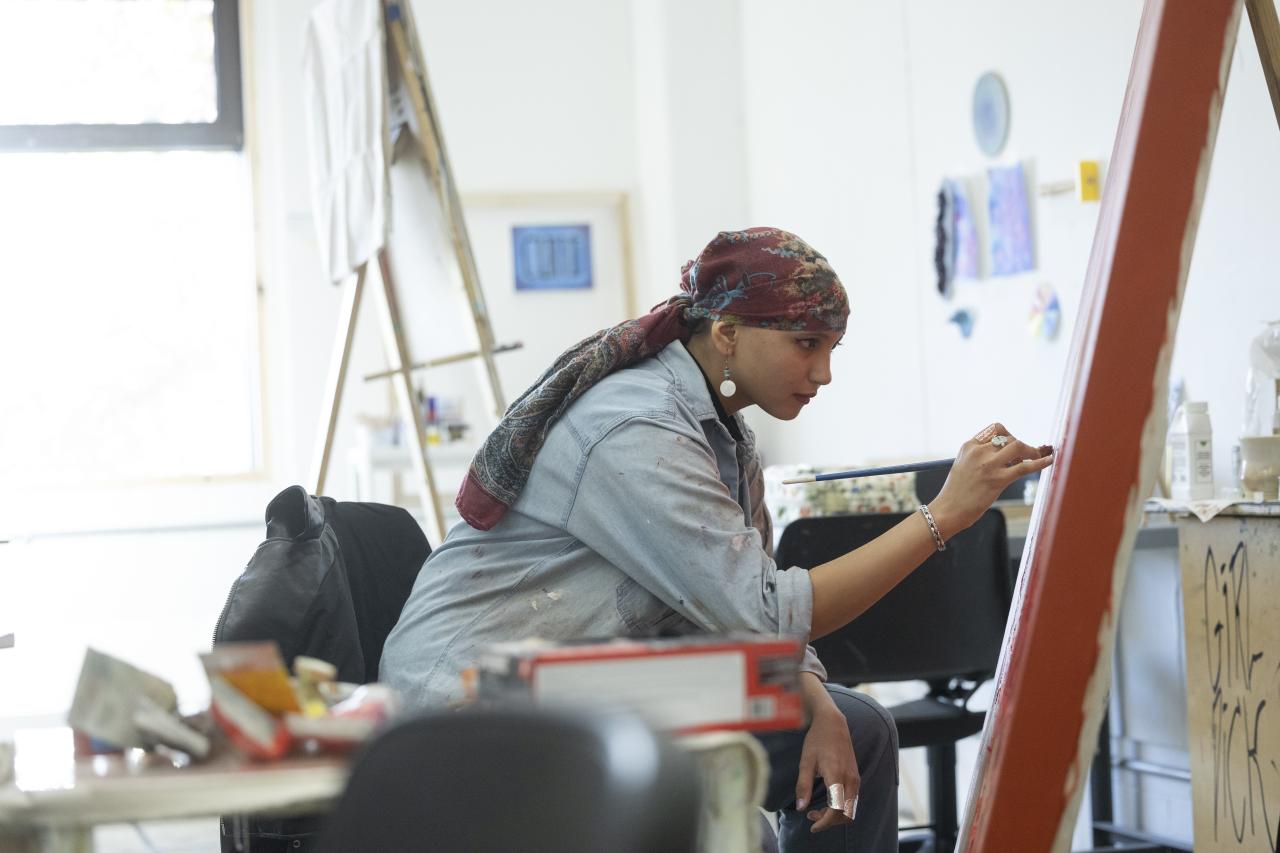 An art student wearing a patterned headscarf and denim shirt sits focused at an easel, carefully painting on a large canvas in a bright studio space.