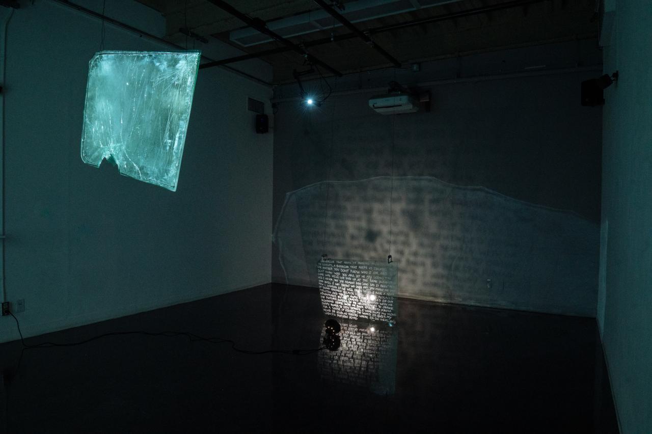 Meghana Iyer, BFA Media Arts Grad, Replaced, Forgotten, and Broken, Windshield, clamps, paint marker and wire. Art installation by Meghana Iyer featuring suspended broken windshields with painted text, illuminated to cast glowing reflections and shadows on the gallery wall and floor.