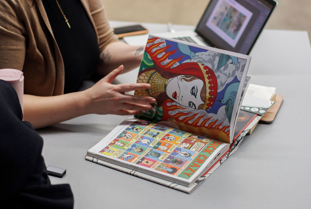 A person points to colorful illustrations in an open comic book, showing a page with a stylized woman surrounded by flames and birds, and another page with a grid of cartoon panels.