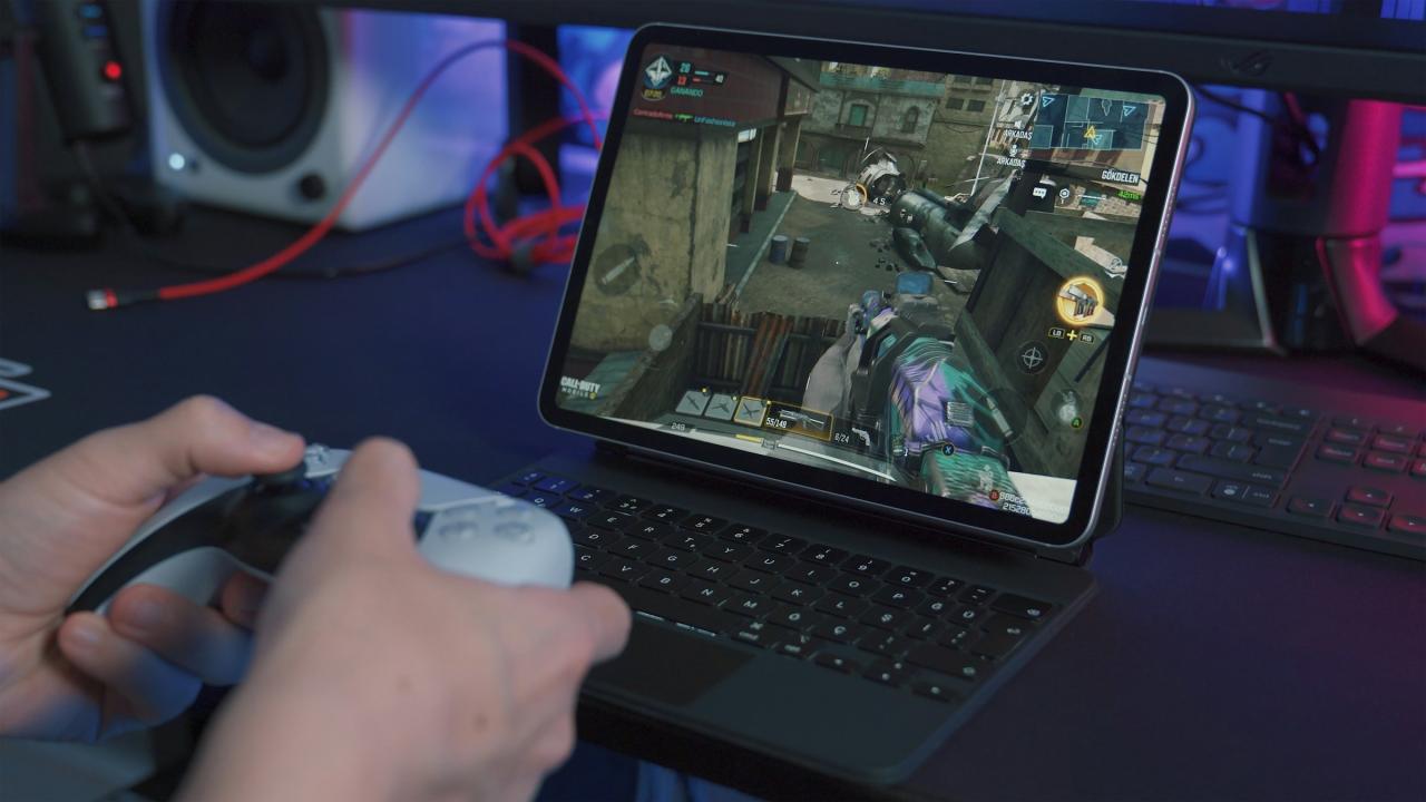 Close-up of a person holding a game controller while playing a first-person shooter game on a tablet. The screen shows colorful graphics of a character in combat, with the tablet placed on a keyboard in a gaming setup.