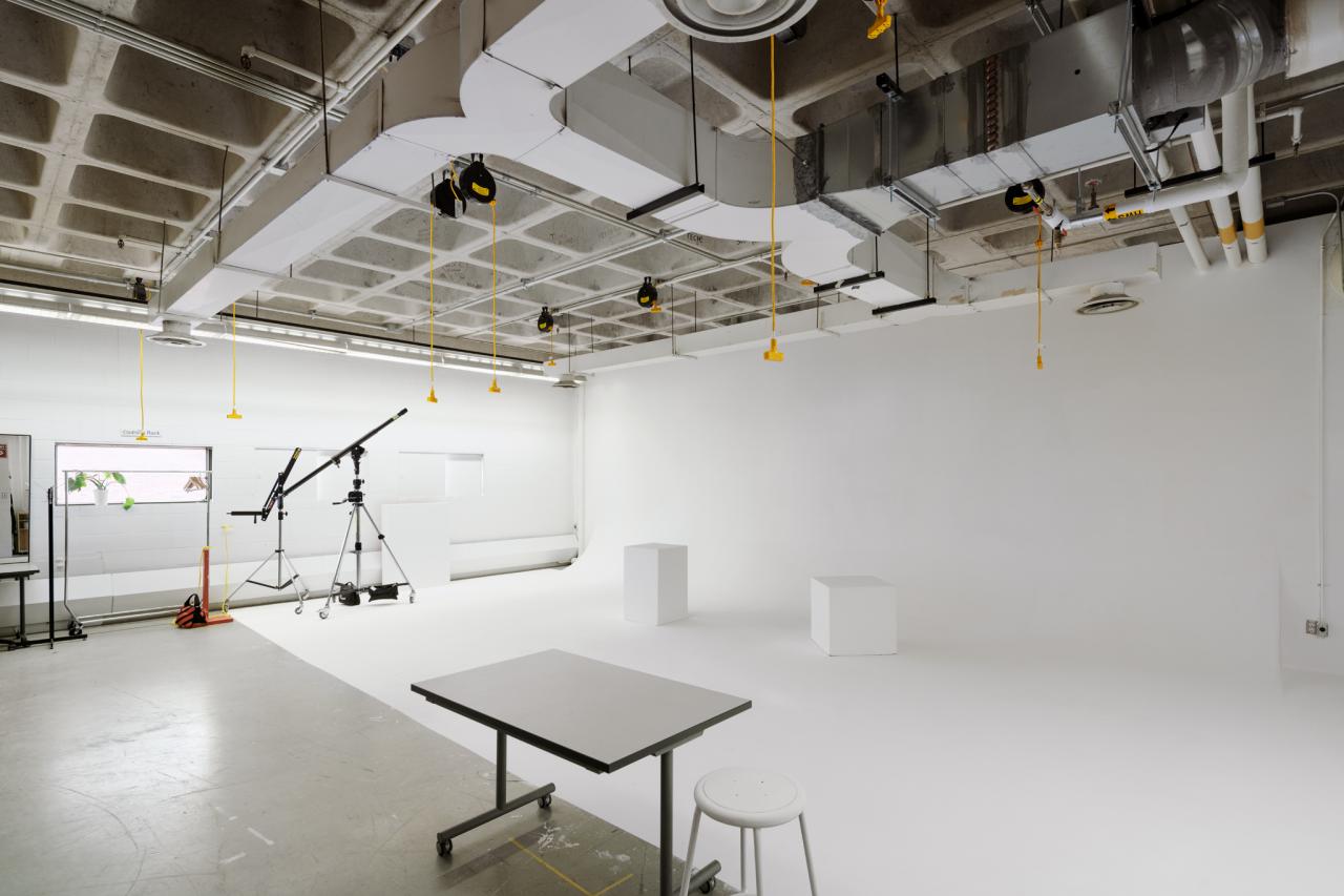 Photo Studio 1 A bright white photo studio with seamless white walls and floor, overhead lighting, and equipment including tripods, a crane arm, and stools.