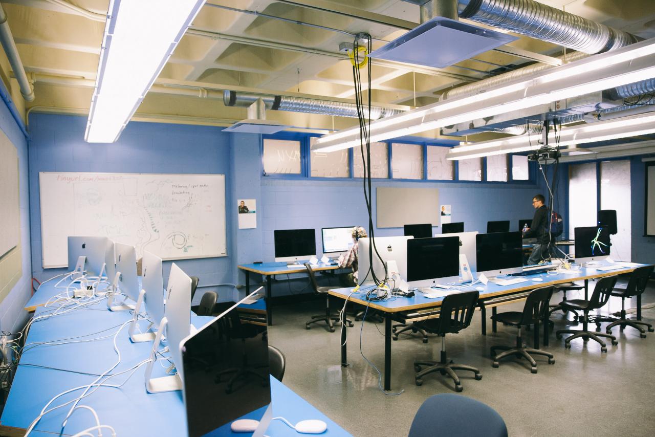 A computer lab with rows of iMacs on blue desks, chairs neatly arranged, and cables running across the tables. A whiteboard with notes and diagrams is visible on the back wall, and a person works at one of the computers while another stands nearby. The room is lit with bright overhead fluorescent lights and exposed ductwork.