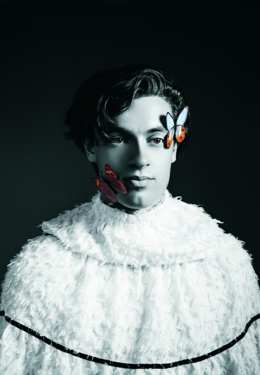 Winner Aigbogun, BDes Photography Grad, This is Us. Black and white portrait of a person in a textured white garment with two colorful butterflies on their face.