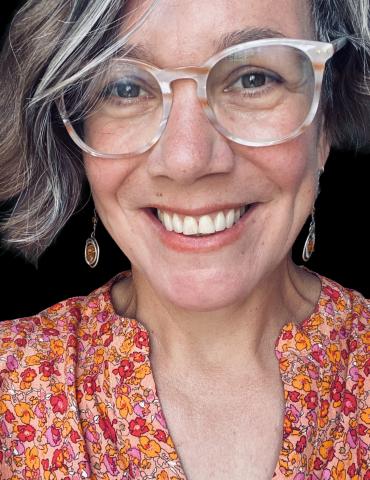 A smiling woman with short, wavy brown and gray hair wears large, clear-framed glasses and dangling earrings. She is dressed in a bright, floral-patterned blouse with orange, pink, and yellow colors. The background is dark, making her face and colorful clothing stand out.