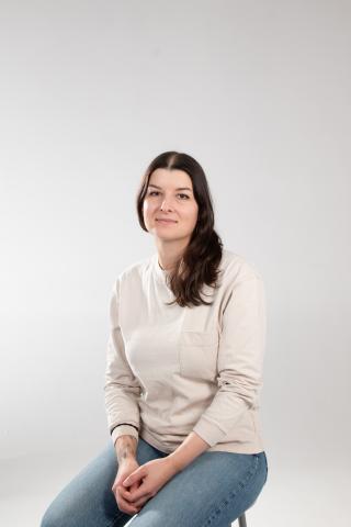 A seated studio portrait of a woman wearing a light beige long-sleeve shirt and blue jeans, photographed against a plain white background. She faces the camera with a calm expression, hands resting together, and her dark hair worn loose over one shoulder.