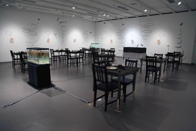 Wide shot of the gallery. We can see the crowded fish tank, tables with whittled down legs occasionally piercing a hot dog. There is a breakfast scene on top each table. There is a fish tank of milk in the far corner. On the walls we see swallow bird vinyl swooping up and down. Between each swallow is a carrot and plate hung on the wall each with their own vinyl thought bubble. 