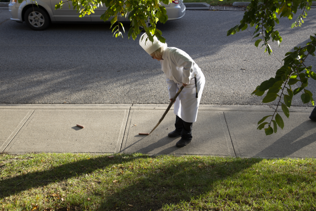 The artist is rolling the hot dogs with a whittled wooden stick down the sidewalk. 