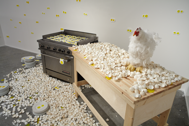 Through the kitchen doors we see the stove top covered in double eggs along with an egg draped over the stove handle. The floor is littered with broken egg shells. Further from the stove there are stacks of plates with the double eggs. There is a raw plywood table with a stressed chicken sitting on top also surrounded by broken egg shells. On the walls there are the double eggs hung around.  
