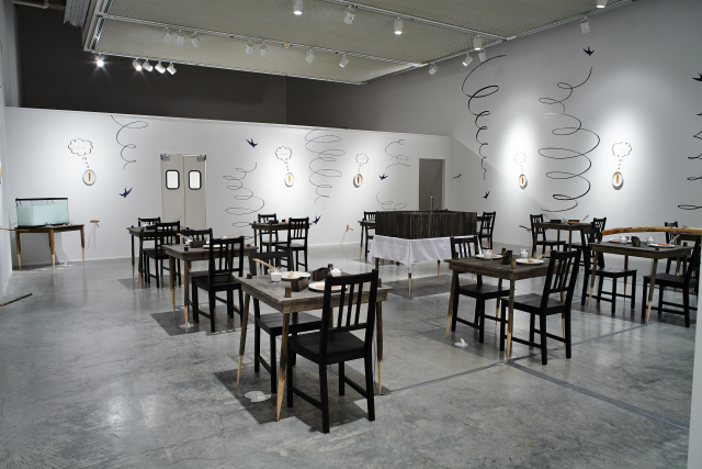 Wide shot of the gallery. From this angle we can see a door leading to another part of the gallery and a wooden cage on a table with a white tablecloth. Tables all have with whittled down legs occasionally piercing a hot dog. There is a breakfast scene on top the tables. There is a fish tank of milk in the far corner. On the walls we see swallow bird vinyl swooping up and down. Between each swallow is a carrot and plate hung on the wall each with their own vinyl thought bubble. 