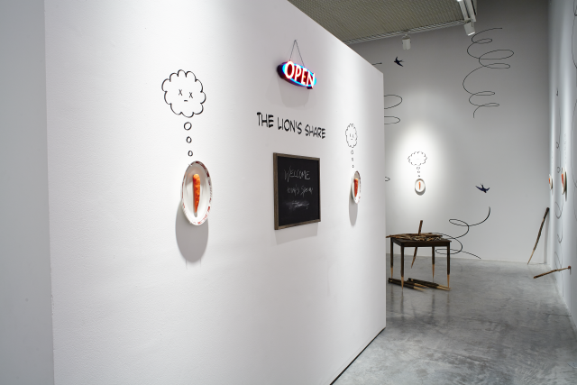 Entering the gallery space, there is a wall that forms a small "waiting section" like a restaurant. There is a generic glowing open sign, under that there is the exhibition title in a comic book font.  A chalkboard hangs under that with the words "Welcome, today's special" and the rest has been smudged. There is a carrot on a plate hanging on each side with an emoji with x's for eyes coming from their vinyl thought bubble. 