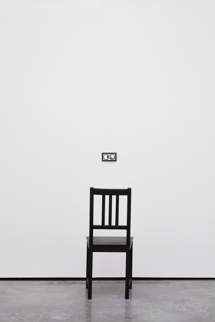 Detail shot of the chair and peephole. The chair is black with a grey cushion. The black rectangle motif mimics the frame around the peephole. From this angle we can start to see through the peep hole. It has glass lenses.  