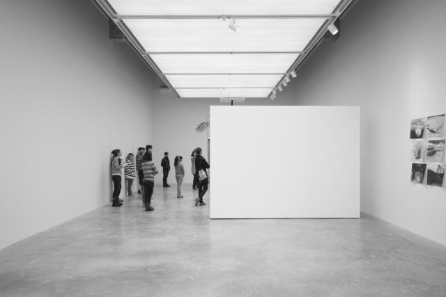 Wide angel of the gallery. There is a floating wall blocking some of the work. We can see the charcoal drawings and a group of people viewing the work behind the floating wall. 