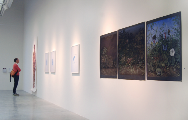 In this image we can see one of the figures with a gallery visitor observing the image. Three of the blue drawings and the pastel triptych are visible as well. 