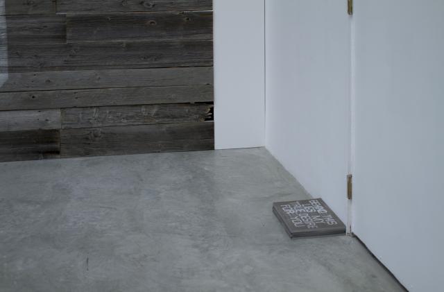 Outside the wall there is a block of wood with the same text as the wall. It is placed in front of a closed gallery door. 