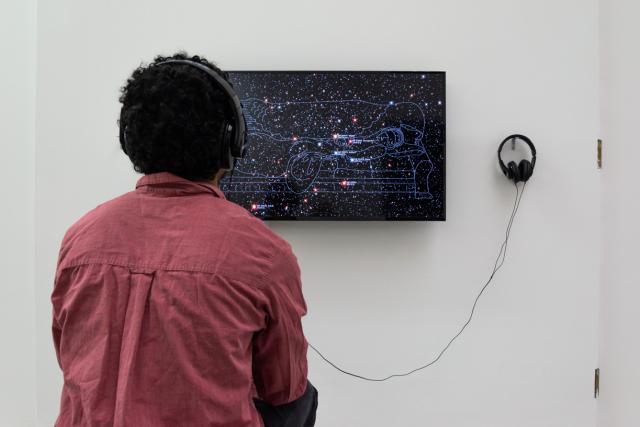 Man watching video on a TV with headphones on, the video shows constellations with a person lying on a recliner like a patient