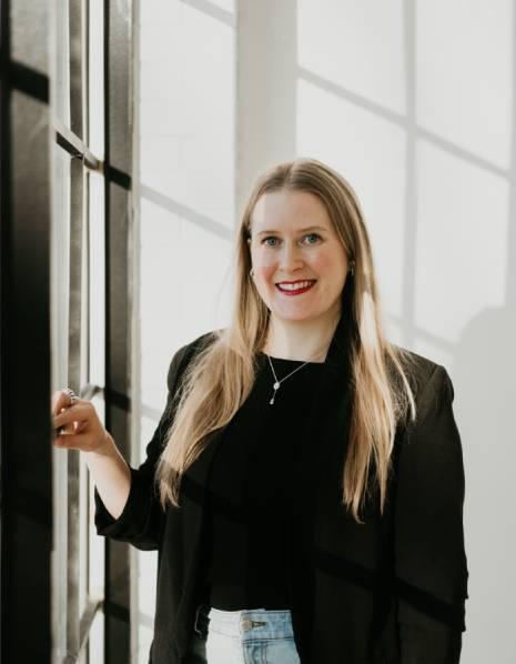 photo of instructor, Kelsey Merkeley, standing in front of a window in a black blazer. 