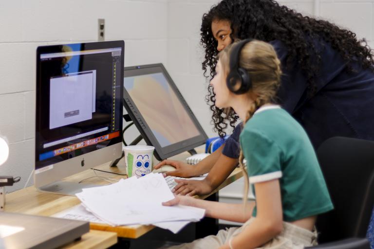 teen in green shirt working at a computer with an instructor assisting them. 