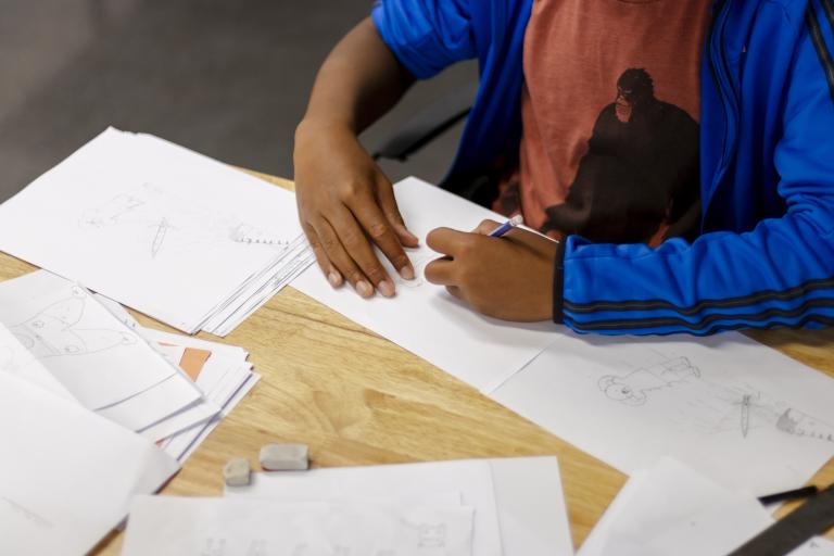 Teen manga student's arm as they work on a drawing project at a table covered in paper and drawings