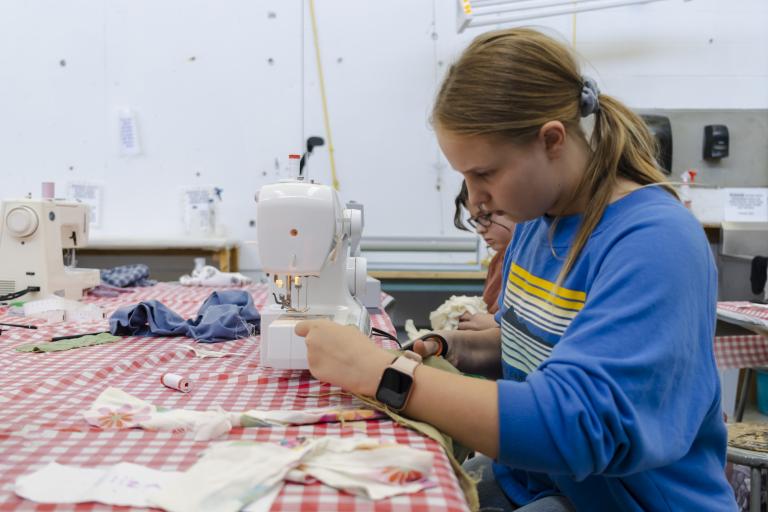 Teen fashion student works at a table with other student using a sewing machine