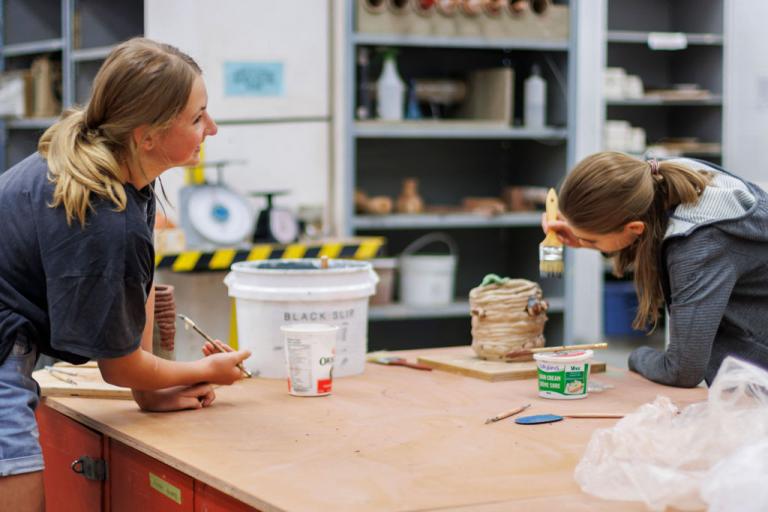 Two teen ceramics students working on hand molded projects at a table smiling.
