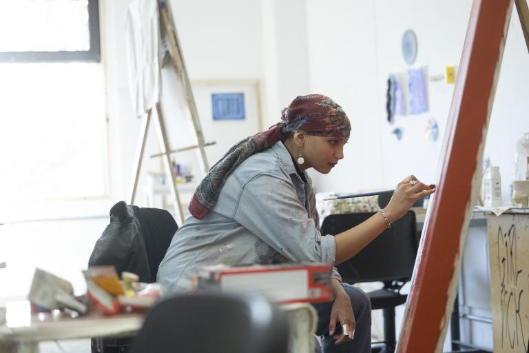 A young artist wearing a patterned headscarf, dangling earrings, and a paint-splattered denim shirt is intently painting on a large red canvas. They hold a paintbrush with precision, leaning in as they apply details. The studio space is filled with easels, artwork, and various art supplies, reflecting a creative environment.