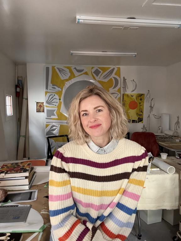 Portrait of artist and AUArts' instructor, Ashleigh Bartlett, in a well-lit studio, wearing a colorful striped sweater and smiling warmly. Behind her, abstract artworks with bold yellow and gray geometric designs decorate the space, alongside creative tools and materials, capturing an inspiring artistic environment.