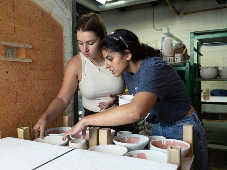 Two students engaged in a ceramics studio, carefully examining freshly fired pottery pieces. The industrial workspace features kilns, shelves of clay works, and tools, creating a hands-on learning environment for ceramic arts.