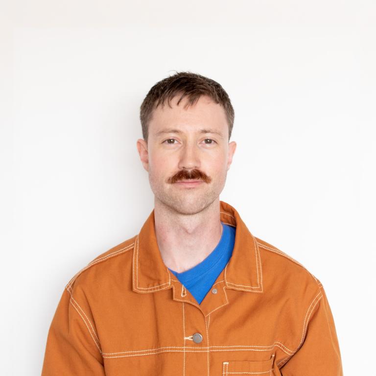 A portrait of Brent Morley Smith, a man with short brown hair and a mustache, wearing a rust-colored work jacket over a blue shirt. He stands against a plain white background, looking directly at the camera with a neutral expression.
