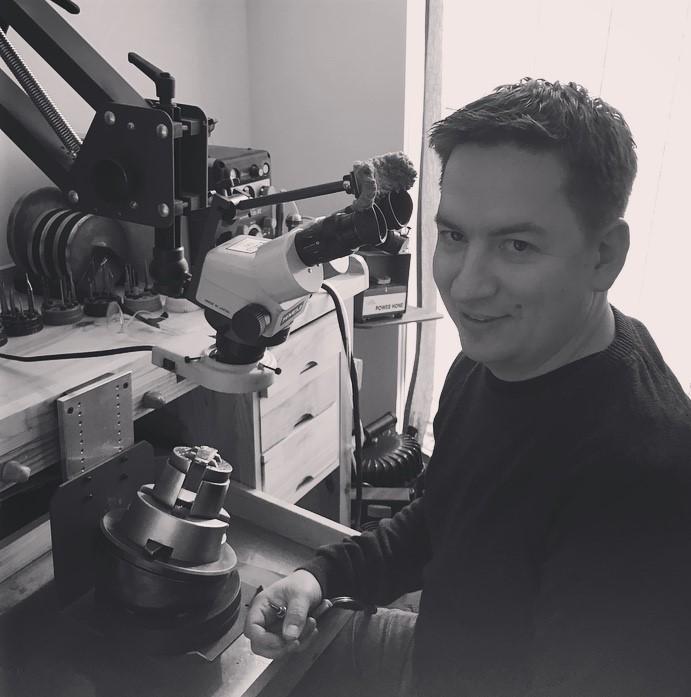 A man with short dark hair and a smile sits at a jeweler’s workbench, holding a tool in his hand. A microscope is mounted in front of him, along with various specialized equipment and tools neatly arranged in the background. The setting is a well-lit workshop, suggesting precision craftsmanship.