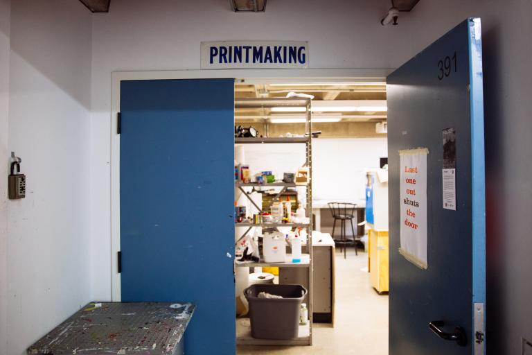 Printmaking studio feature image A blue door labeled “Printmaking” stands open, revealing shelves with inks, tools, and supplies inside a studio. A sign on the door reads, “Last one out shuts the door.