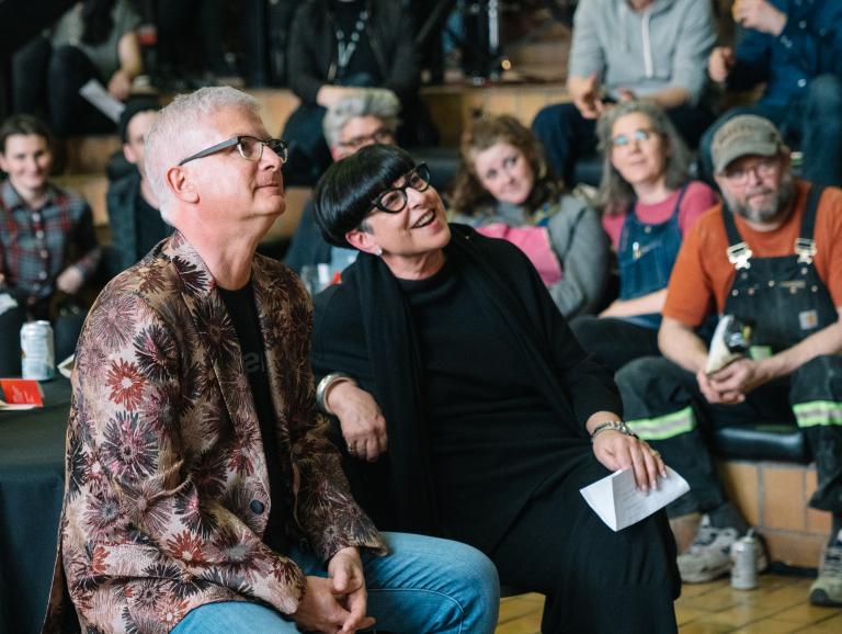 A seated group at an AUArts event smiles and looks toward the stage; two people in the front row lean together, appearing engaged and cheerful.